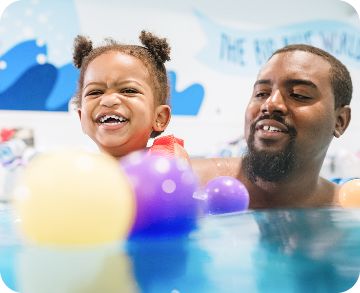 A young child in a colorful swimsuit enjoys playing with floating balls in a swimming pool, with playful wall art in the background.