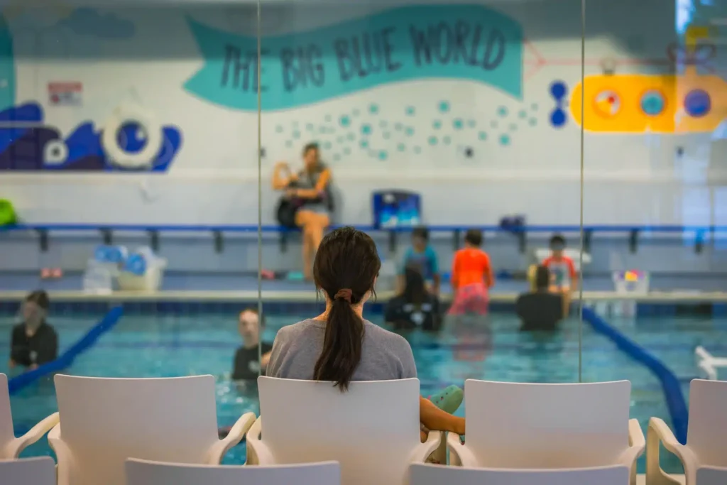 mom sitting in Big Blue waiting area looking out to the pool where swim lessons are happening