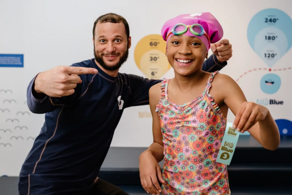 Swim instructor enthusiastically pointing to one of his students who just got a Big Blue ribbon for completing this swim skill