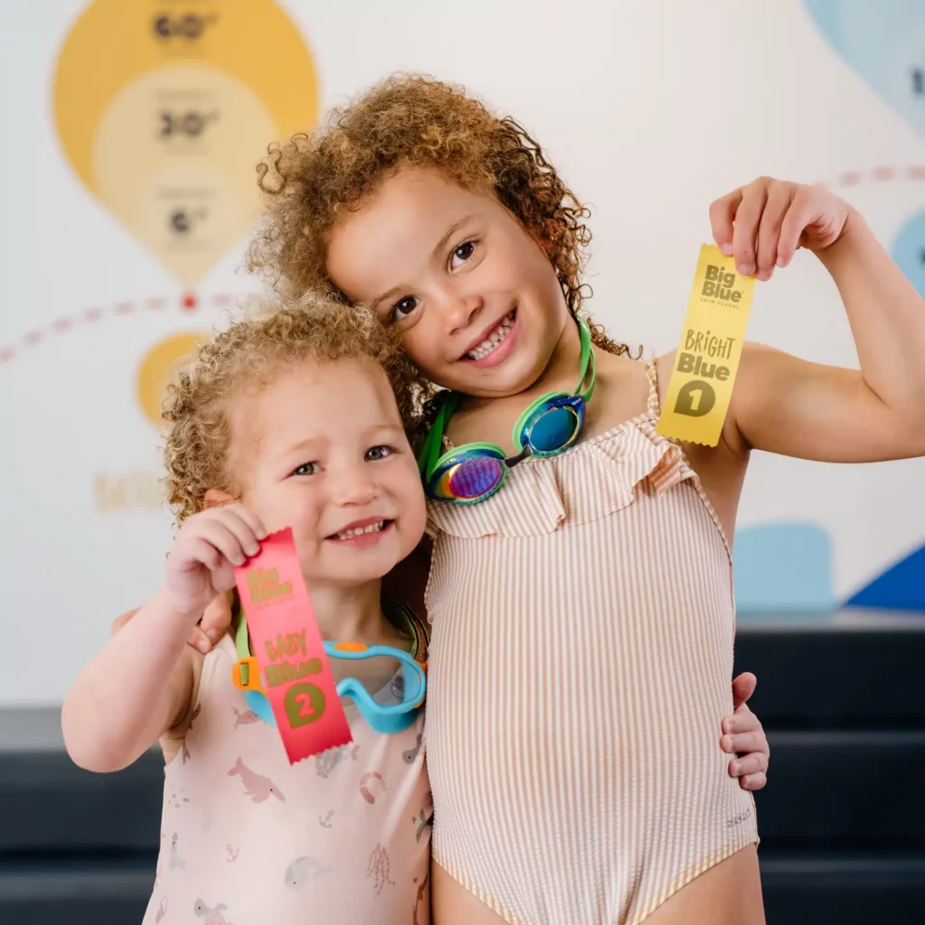 two young girls standing next to each other and hugging. Both are holding swim ribbons and a Big Blue Swim School medal