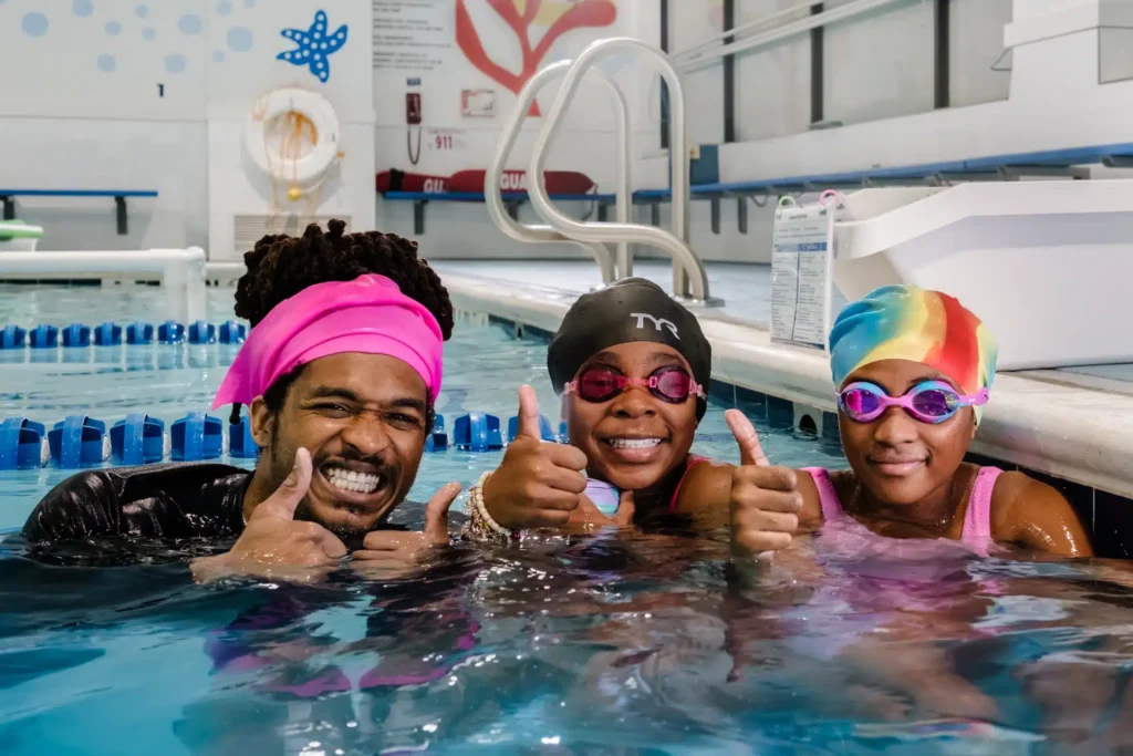 an instructor and two girls in swim caps and goggles smiling at the camera giving a thumbs up