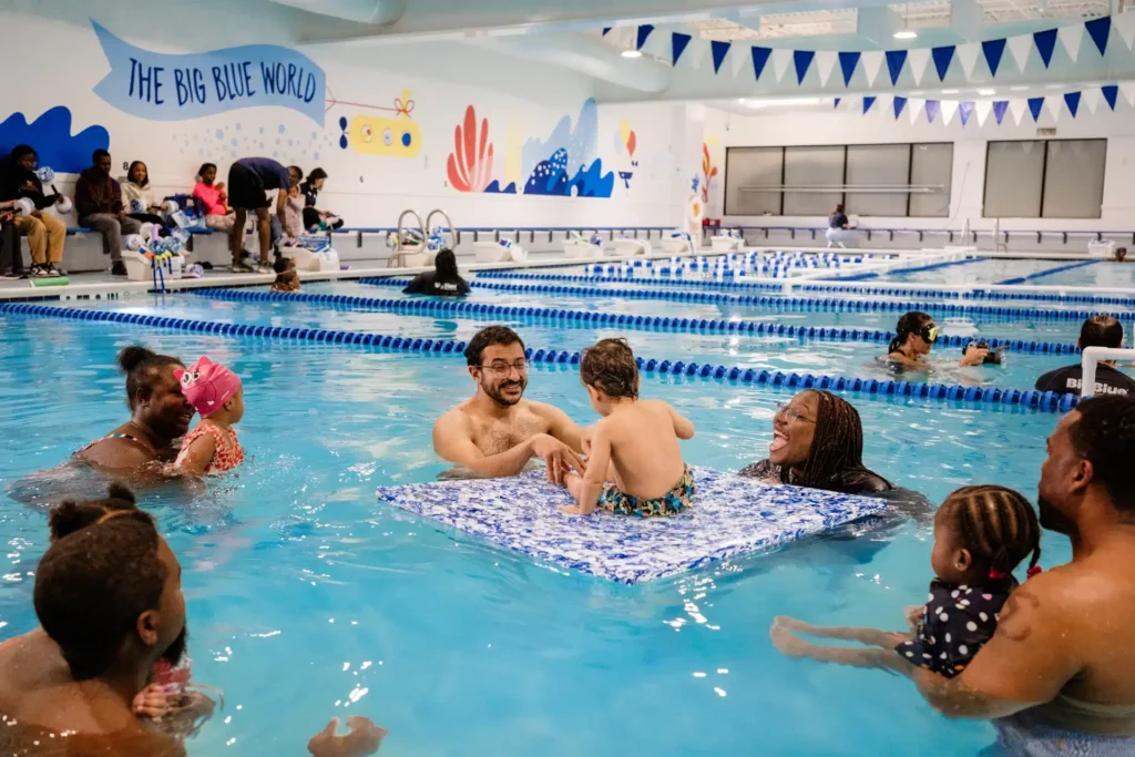 a baby blue swim lesson with parents and children swimming and using a floating mat for the babies to get used to the water