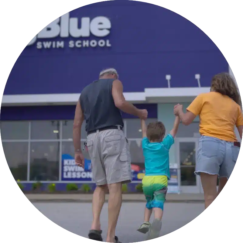 two parents holding a young child's hands, who is in swim trunks, walking towards a Big Blue Swim School