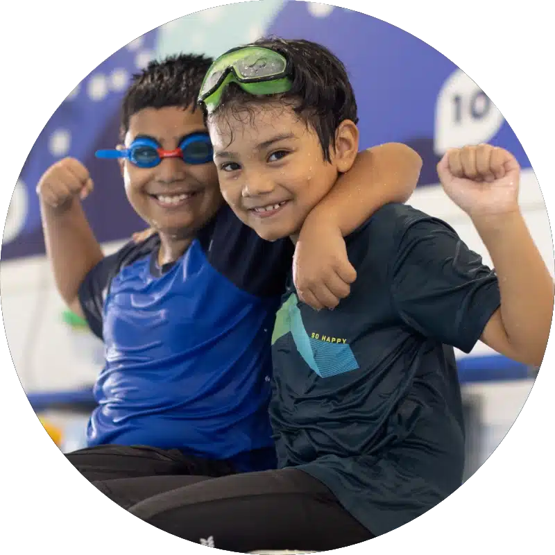 two smiling boys with swim goggles sitting next to each other holding their fists up in triumph