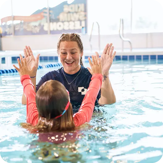 A swim instructor and a young girl in the pool, both with their hands raised in a "high five" gesture, smiling at each other.