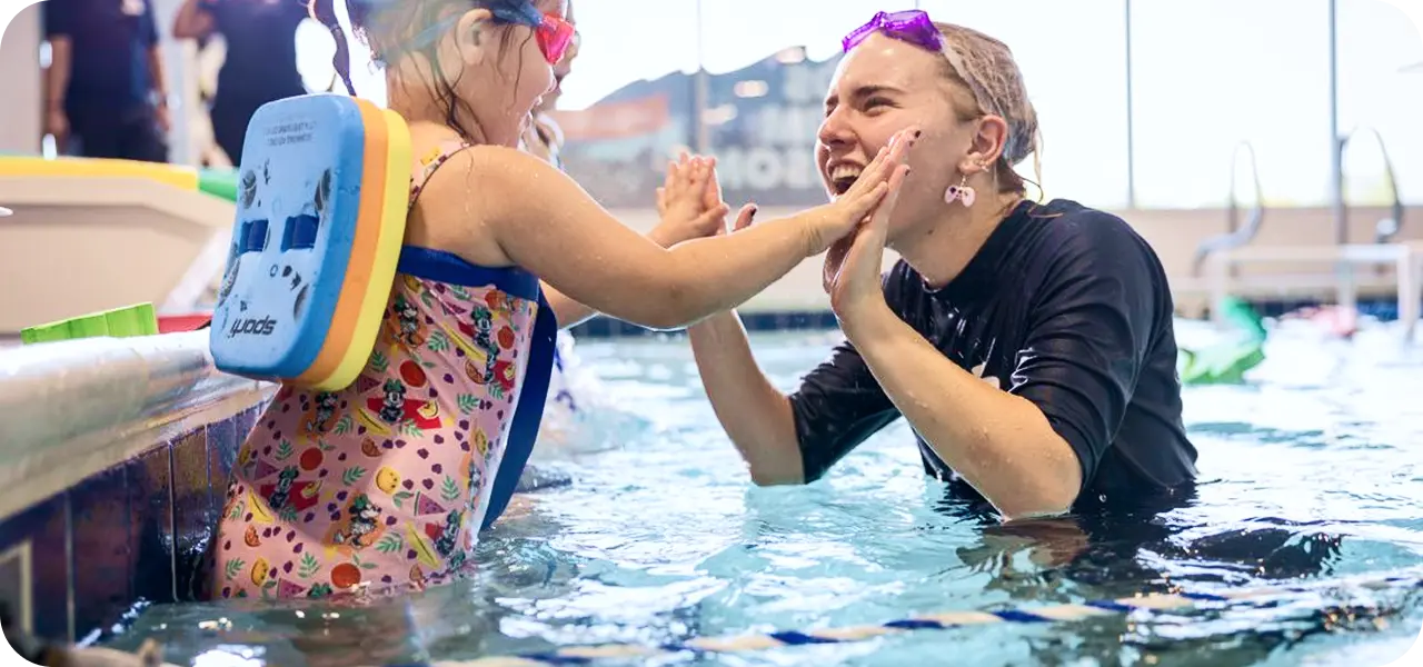 A young girl in a patterned swimsuit and a swim float, wearing pink goggles, high-fives a smiling swim instructor wearing purple goggles and a black swim shirt in a swimming pool.