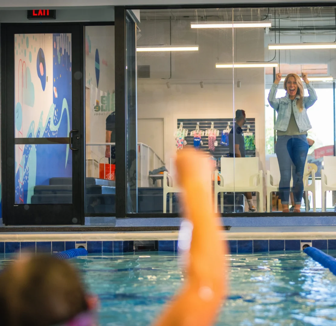 a mom behind the glass in the waiting room listing both hands in support, looking towards a kid in the water who is raising their fist in triumph
