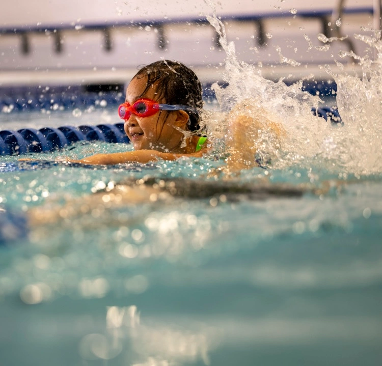 a girl in pink goggles swimming in a pool and splashing the water up