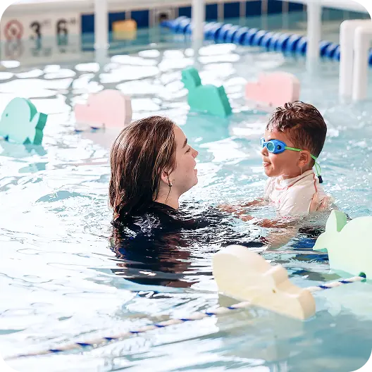 A young child in blue swimming goggles is held in water by an instructor, both smiling at each other in a pool surrounded by various colorful floatation devices.