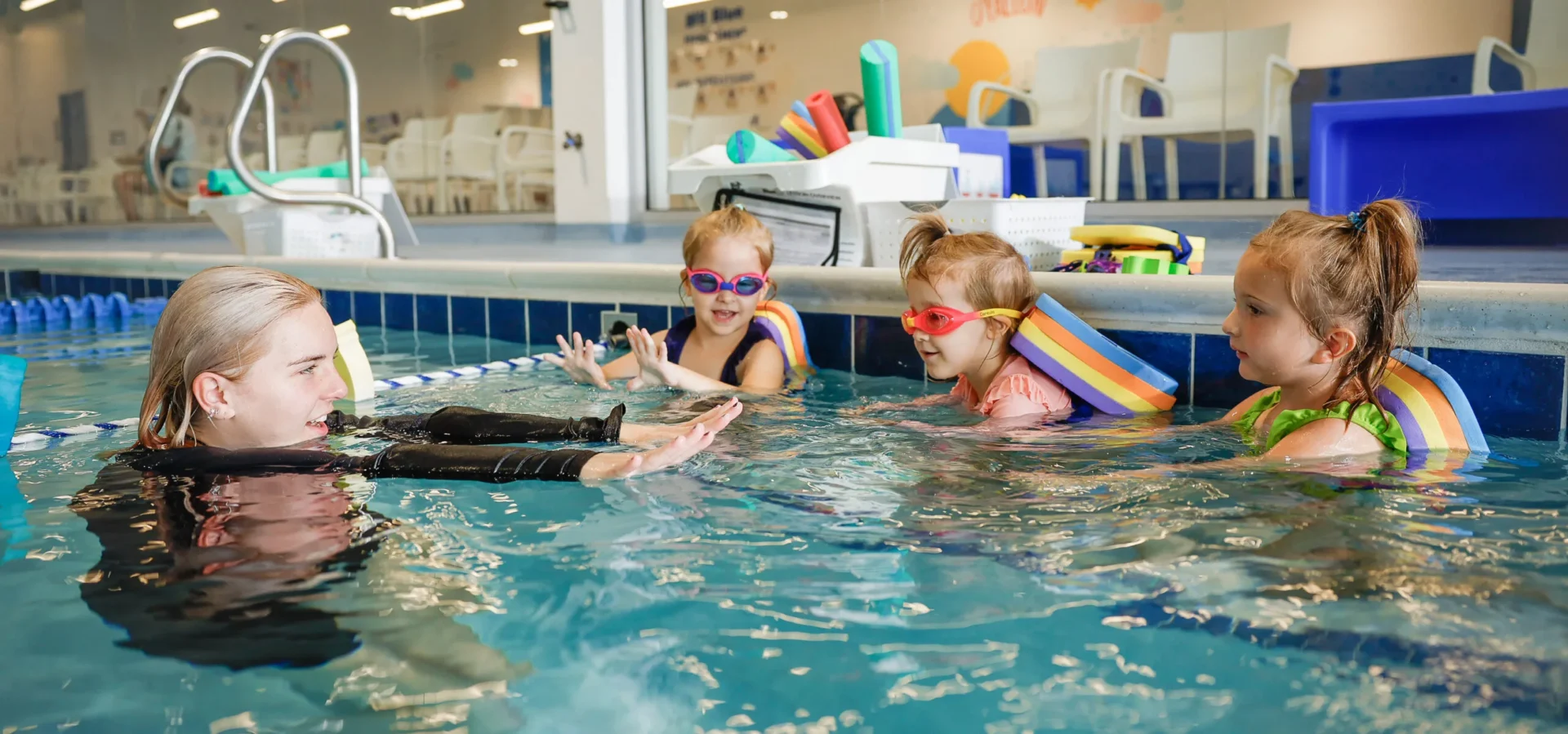 a swimming instructor in a pool giving a demonstration of a swimming stroke to three girls wearing floatations devices and goggles